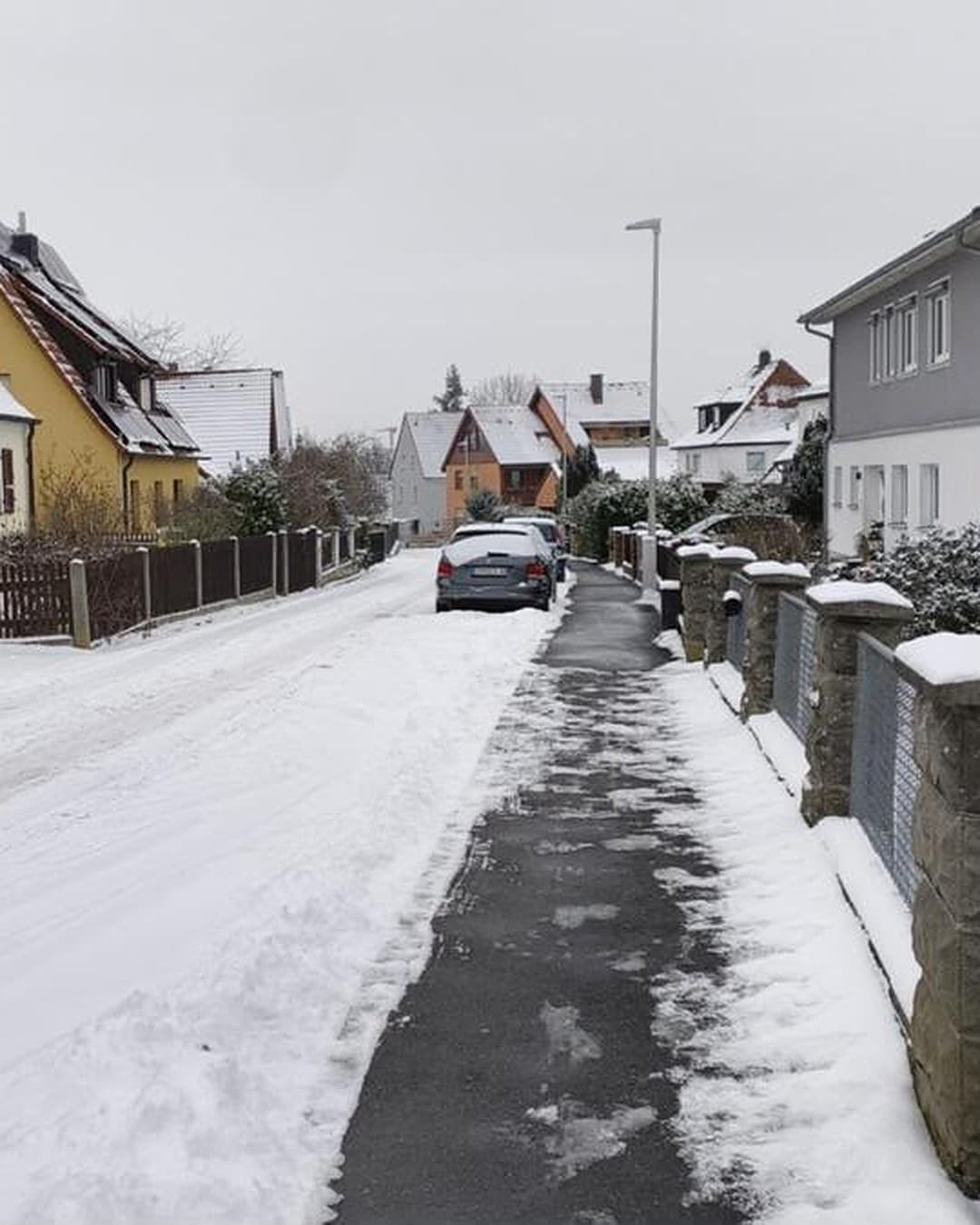 Geräumter Gehweg neben einer verschneiten Wohnstraße, rechts Zaunpfeiler mit Schneehauben